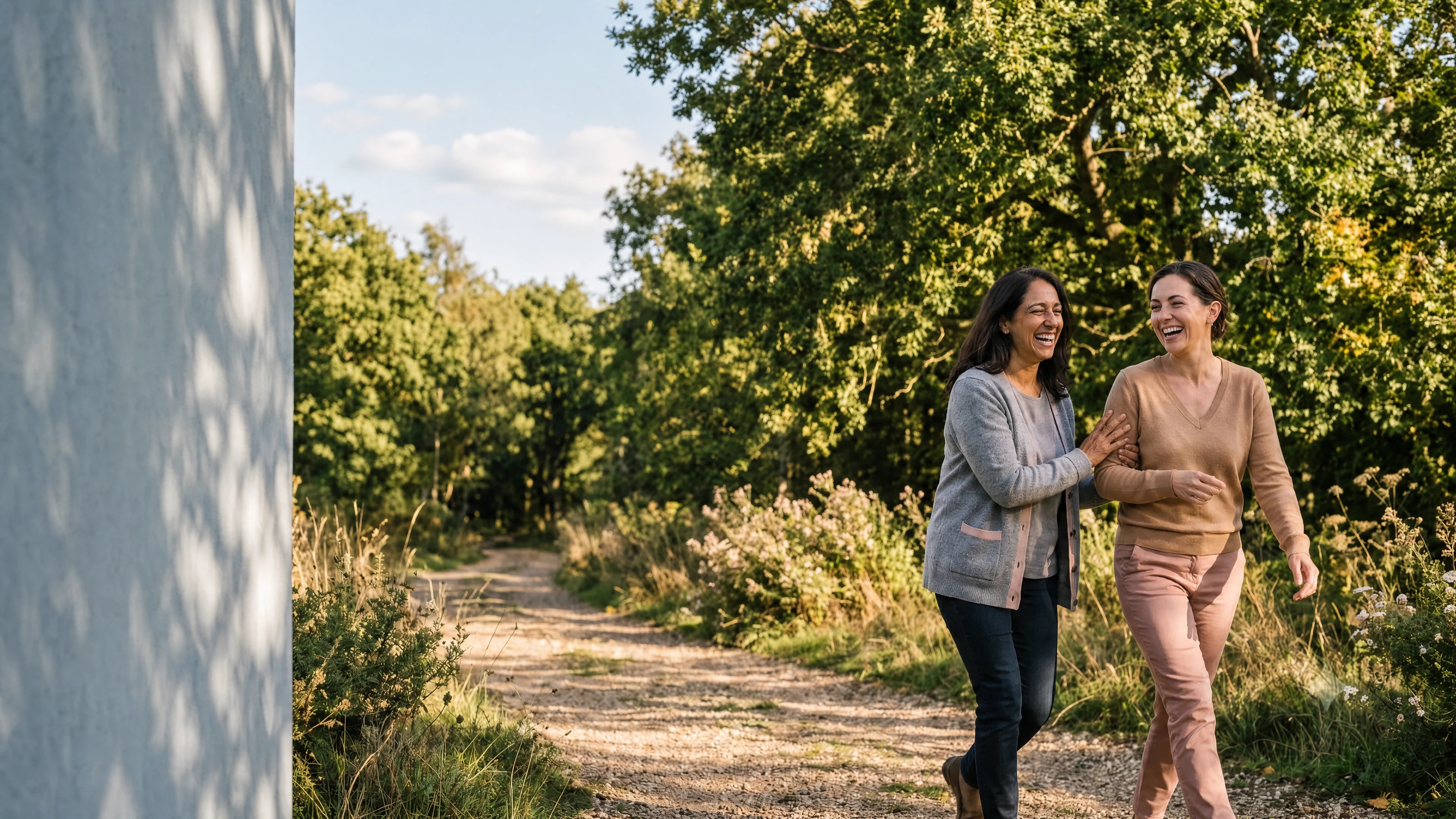 Two women laughing together on a sunlit, tree-lined path